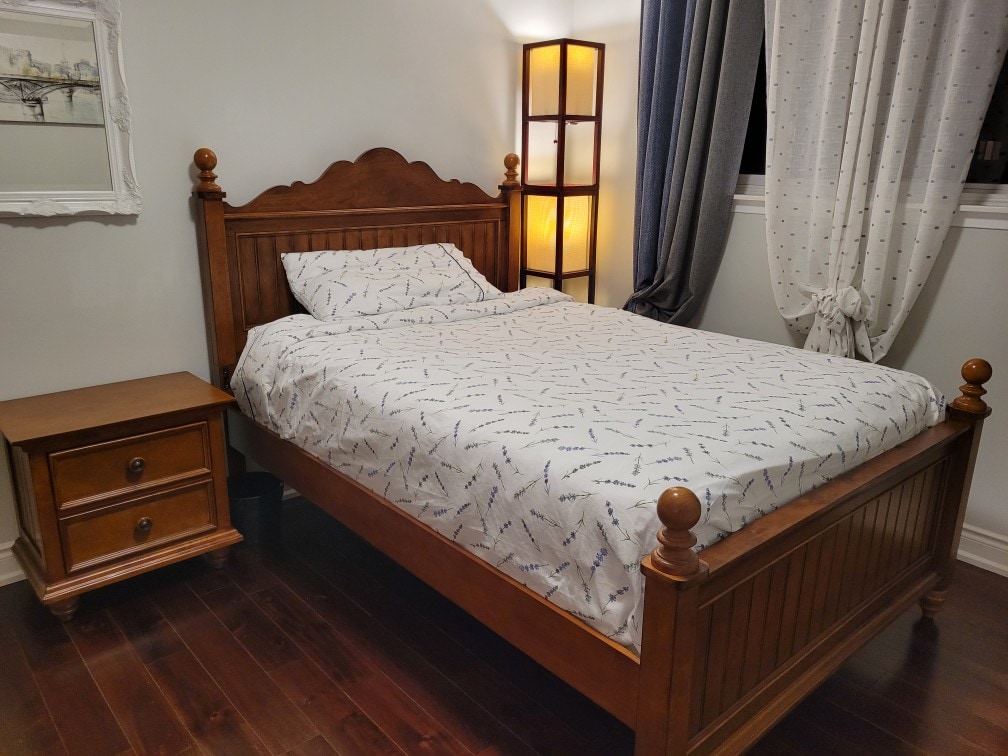 A cozy bedroom features a wooden bed with a patterned white duvet and matching pillows. A small nightstand is located beside the bed, complementing the warm wood tones. Natural light filters through the draped curtains, and a soft lamp illuminates the space.