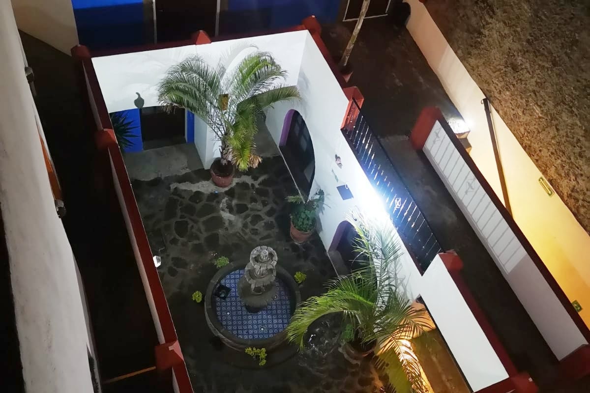 An aerial view reveals a charming courtyard featuring a small stone fountain at its center, surrounded by vibrant greenery and potted palm trees. The walls are painted white with colorful accents, while a blue sky is visible above, creating a serene and inviting outdoor space.