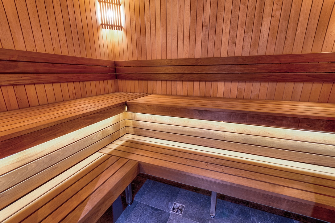 A warm sauna features smooth wooden benches arranged along the walls, creating ample seating space. Soft lighting highlights the natural wood tones, contributing to a relaxing environment. The floor is composed of dark tiles, contrasting with the lighter wooden surfaces.