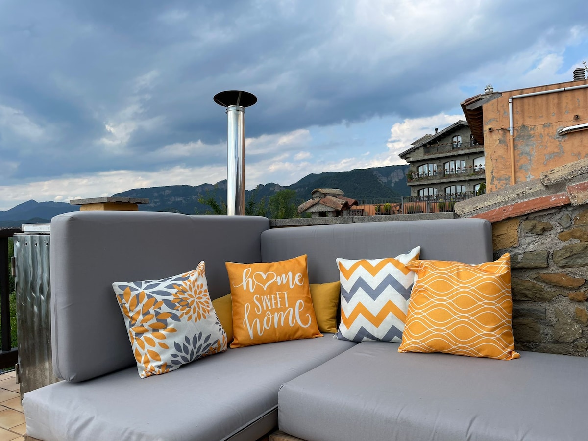 A comfortable outdoor seating area is featured, showcasing a spacious gray sectional adorned with decorative throw pillows in various patterns and warm colors. A scenic view of surrounding hills and buildings adds to the cozy ambiance, complemented by an outdoor heater.