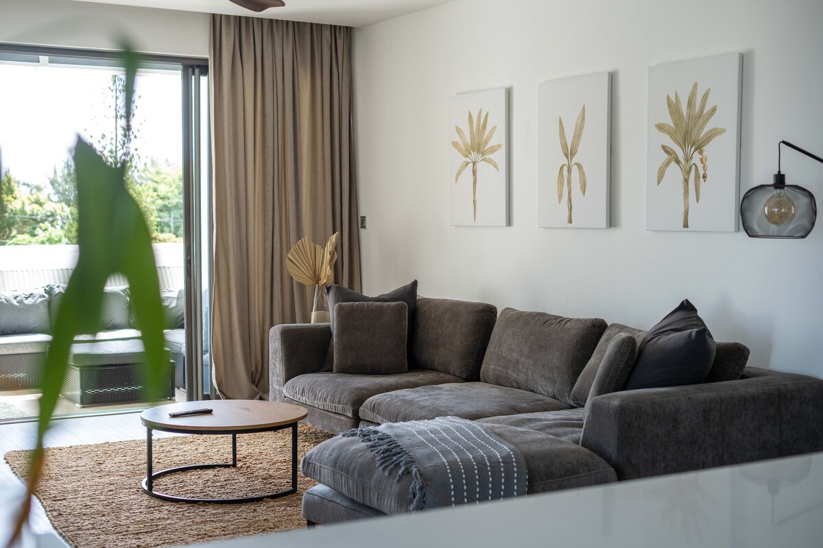 A spacious lounge area features a comfortable gray sectional sofa arranged around a circular coffee table. Three botanical prints adorn the white wall, while a large window with sheer curtains brings in natural light. A woven rug adds warmth to the space.