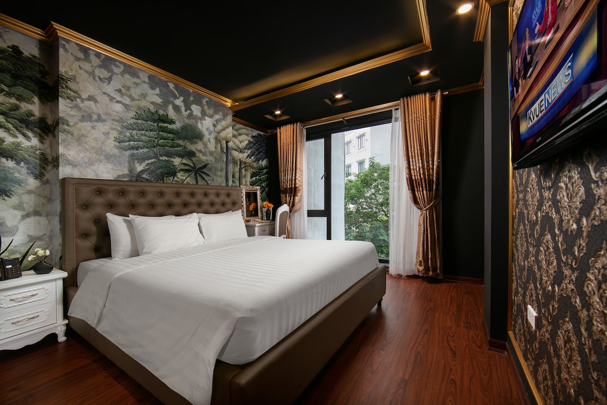 A spacious suite is presented, featuring a tufted bed with crisp white bedding. Elegant wallpaper with a natural motif lines the walls. Large windows allow natural light to brighten the room, while decorative curtains frame the view. A flat-screen television is mounted on the wall.