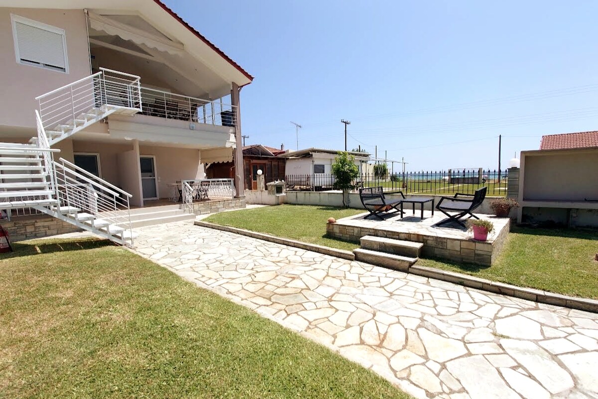 The image displays the exterior of a two-story villa, featuring a stone pathway leading to a grassy area. Outdoor lounge chairs are positioned on a patio. A stairway leads to the upper floor, and the surrounding space is well-maintained, offering an inviting atmosphere.