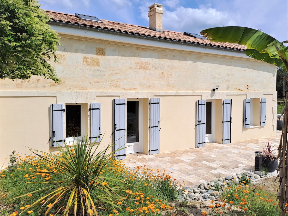 The exterior of the house features a traditional stone façade complemented by blue shutters. Windows are framed to enhance natural light in the interior. An outdoor patio is adorned with flowering plants and landscaped greenery, providing a serene space for relaxation.