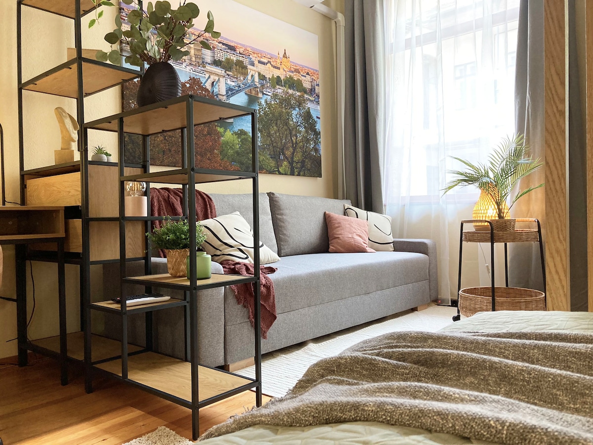 A living area is presented with a comfortable gray sofa and decorative cushions. A shelving unit holds small plants and decorative items, enhancing the space. Natural light filters through large windows, illuminating the warm wood flooring, while a cozy textile covers the bed in the foreground.