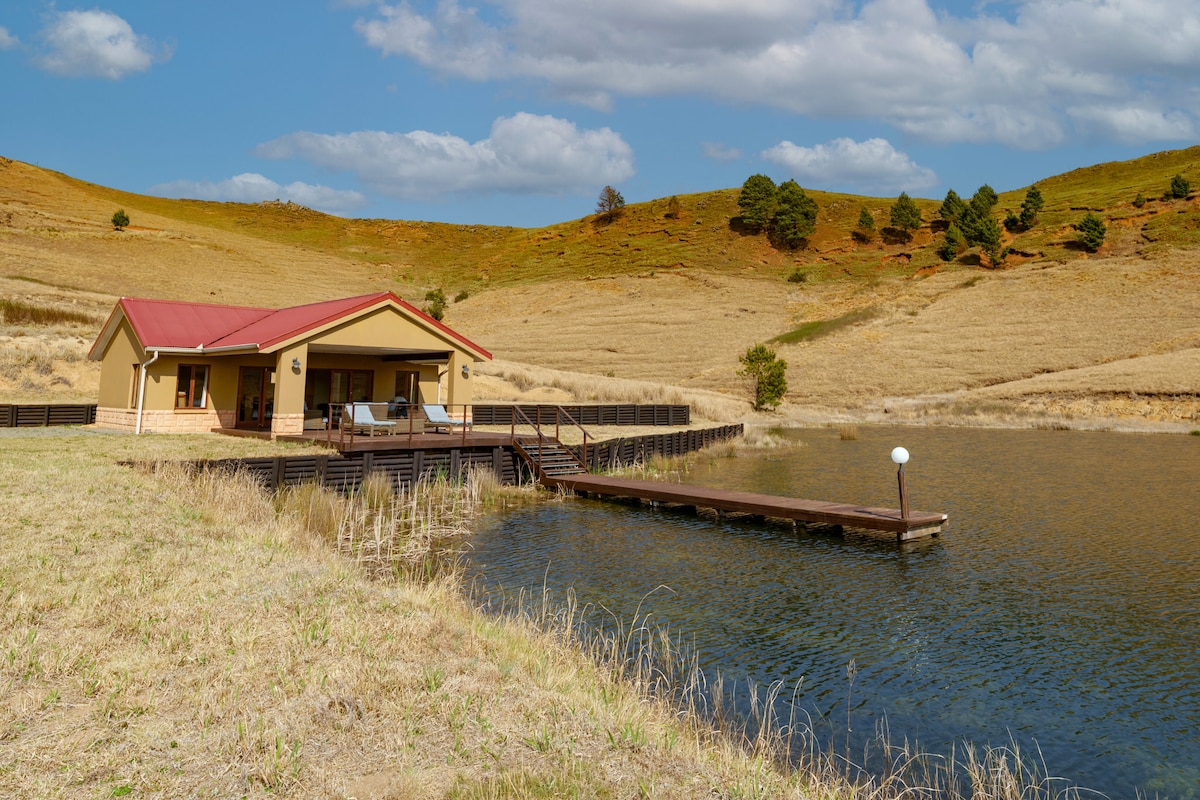 A modern lake house is situated by a serene body of water, surrounded by gently sloping hills. The exterior features a charming red roof and a spacious wooden deck leading to a dock. The landscape includes dry grass and patches of greenery.