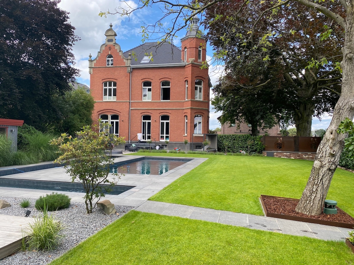 A red brick mansion is surrounded by well-maintained lawns, featuring a modern pool that reflects the sky. Mature trees provide shade, while landscaped areas with pebbles add texture. The building showcases tall windows, with a charming architectural style prominent against the vibrant greenery.