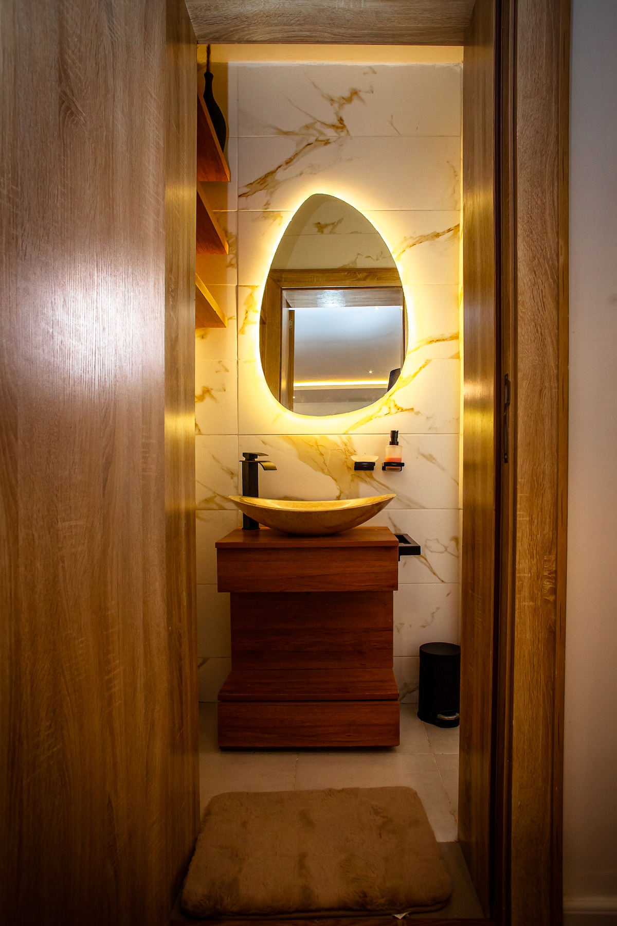 A compact bathroom area is showcased with sleek marble walls and subtle lighting. An oval mirror is illuminated above a modern sink unit made of wood. Shelving is integrated into the wall for added storage, and a small rug is placed in front of the sink.