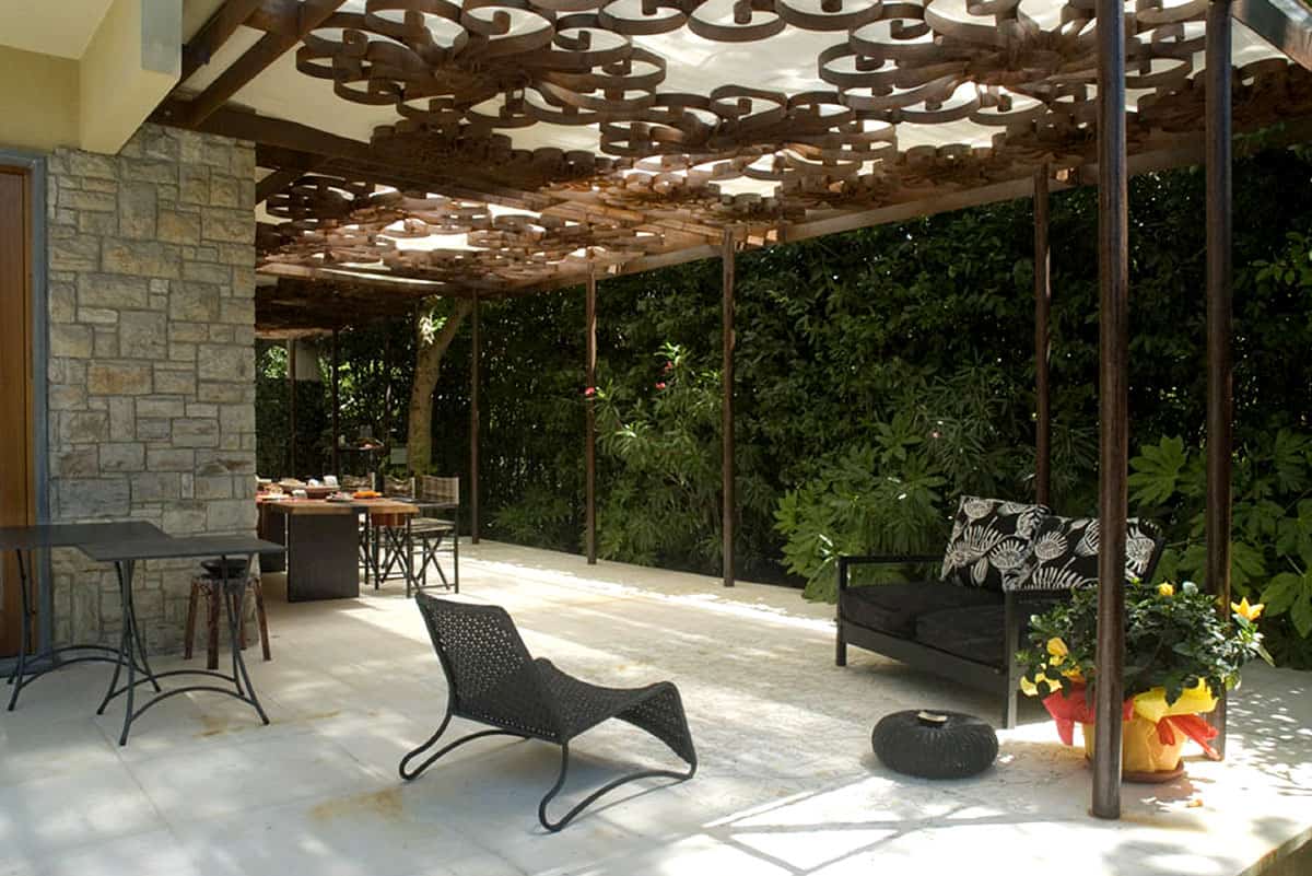 A shaded outdoor seating area is presented, featuring a stylish black lounge chair and a dark sectional sofa. Natural greenery surrounds the space, complemented by a decorative table set for dining under an intricate wooden overhead design.