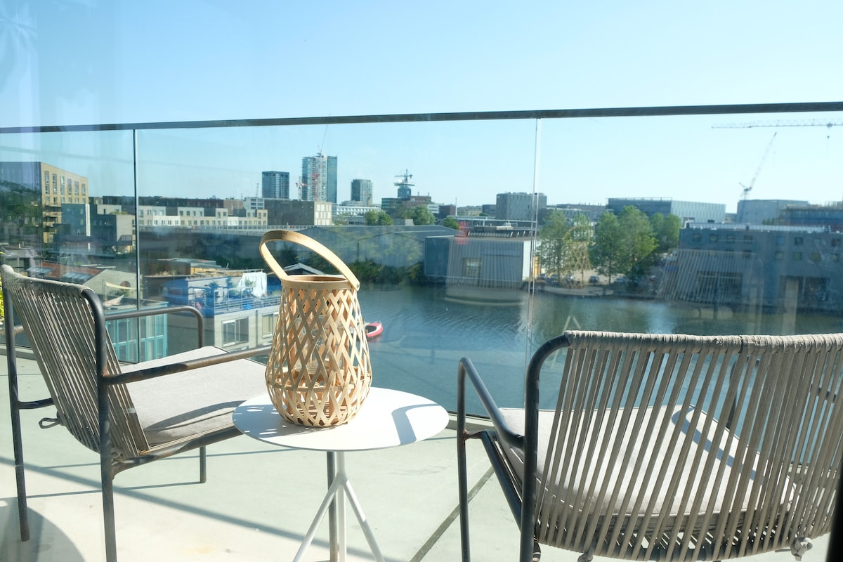 A balcony setting features two elegant chairs beside a round table. A woven lantern adds a decorative touch, with a scenic view of the water and modern buildings in the distance. Clear glass railings provide an unobstructed sight of the surroundings.