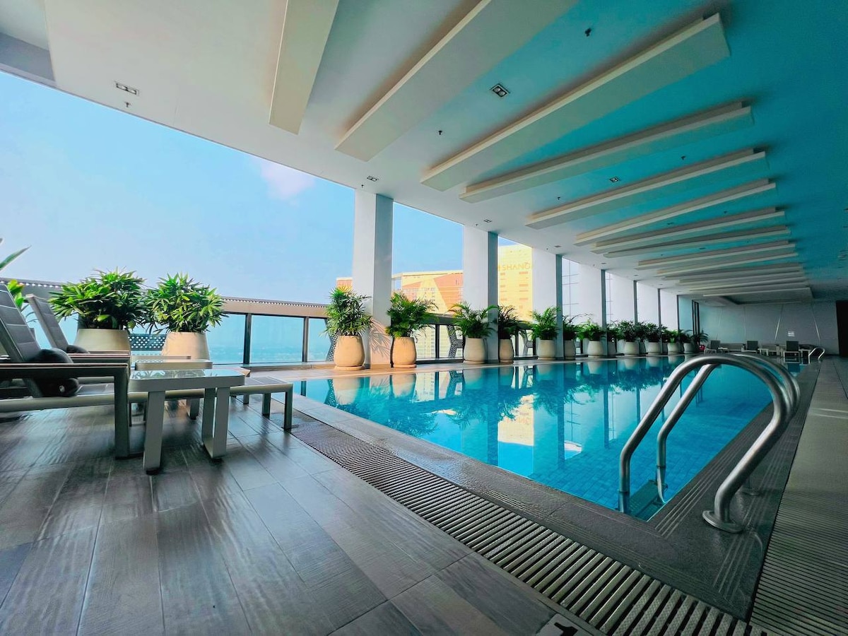 An indoor swimming pool is framed by large windows that allow natural light to fill the space. Potted plants line the edges of the pool area, while loungers are positioned nearby. The flooring is tiled, providing a sleek, modern appearance.