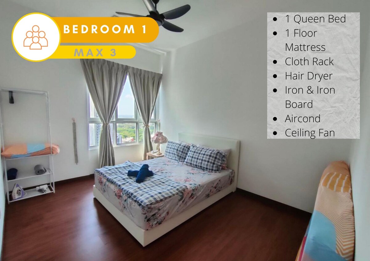 A bright and spacious bedroom features a queen bed with a patterned blanket and a nearby floor mattress. Natural light fills the room through large windows dressed with drapes. A cloth rack and a hair dryer are also visible, along with an iron and ironing board.