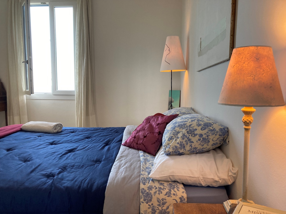 A spacious bedroom is highlighted by a comfortable bed adorned with patterned linens and decorative pillows. Soft light is emitted from two bedside lamps, creating a tranquil ambiance. A window is partially draped with light curtains, allowing natural light to fill the room.