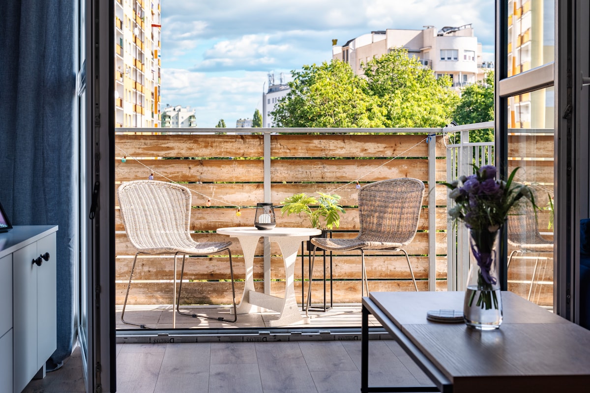 A balcony is depicted with two woven chairs and a small table, providing a space to relax outdoors. A potted plant sits on the table, and greenery from nearby trees can be seen in the background, complementing the urban view.