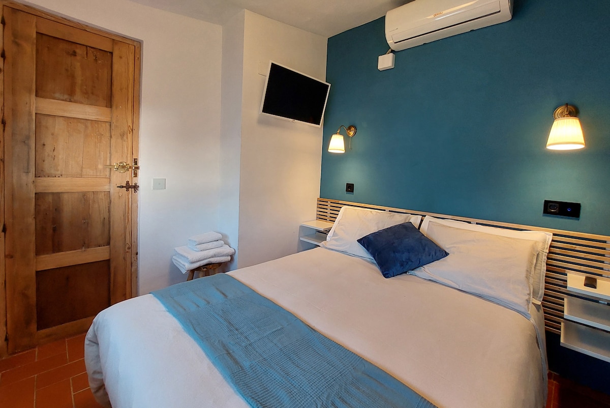 A room is presented with a spacious bed featuring white linens and a blue accent pillow. Wall-mounted lights provide soft illumination next to the bed. A wooden door leads to the hallway, and a flat-screen TV is mounted on the wall opposite the bed.