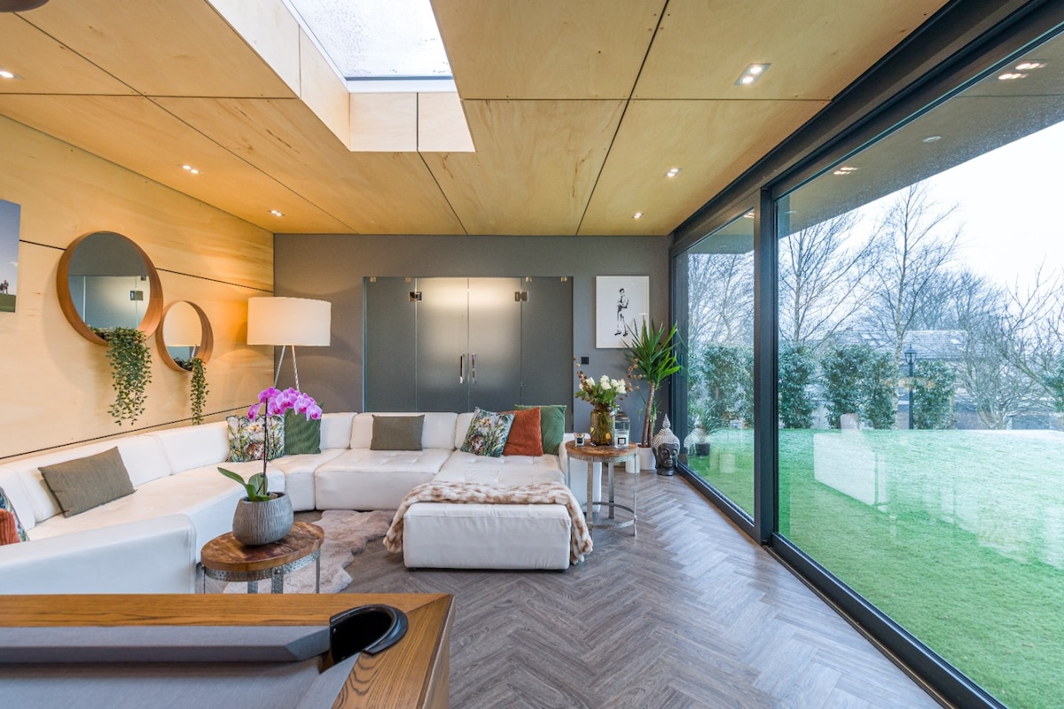 A spacious open-plan living area features a curved sectional sofa adorned with warm textures. Natural light floods in through large glass walls, offering views of the garden. A wooden table holds potted plants and decorative items, complementing the calming interior design.