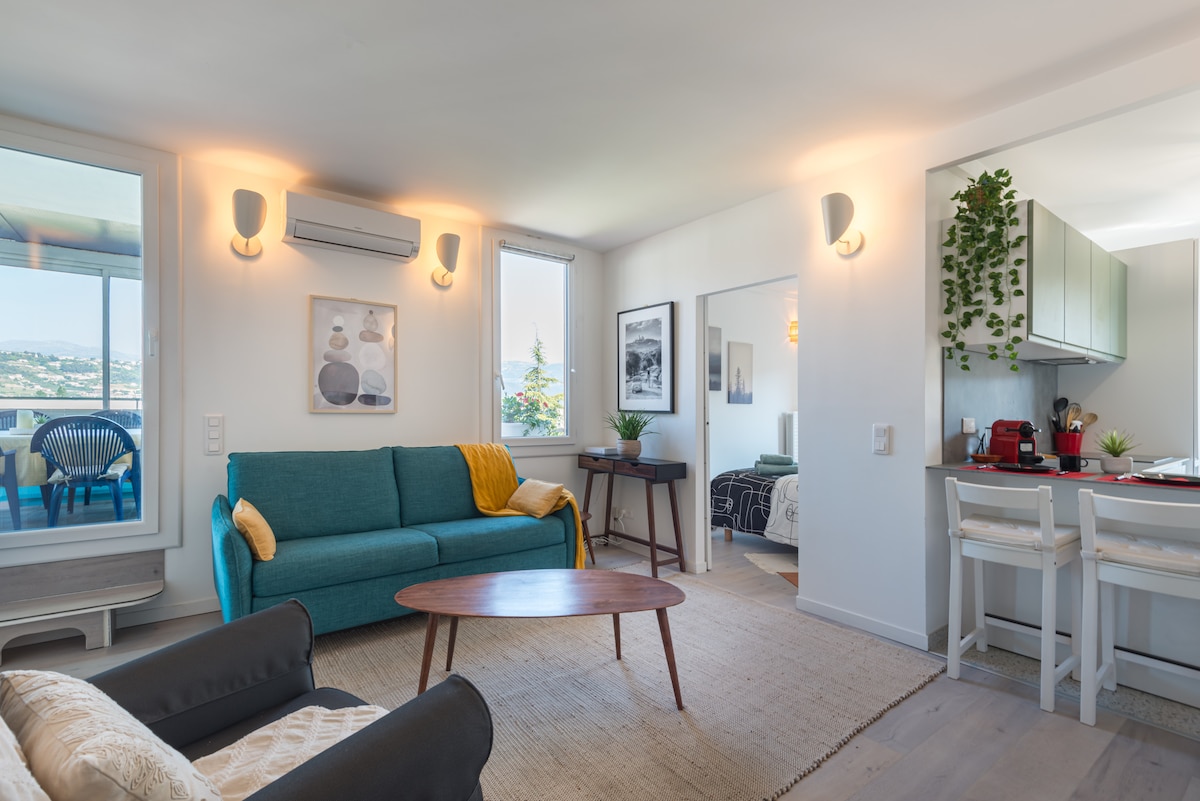 A modern living area features a teal sofa and a round wooden coffee table. Natural light floods in through large windows, offering views of the surroundings. A compact kitchen area with contemporary cabinetry is visible in the background, complemented by minimalist decor.