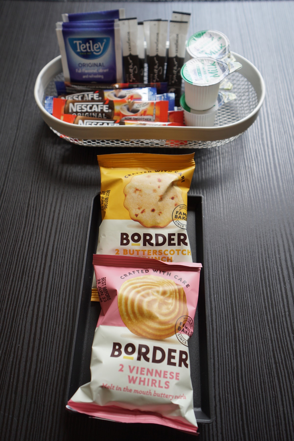 A selection of tea and coffee products is arranged in a basket, featuring various sachets and a few packets of sweeteners. Below, two packets of Border biscuits are displayed, labeled as butterscotch and Viennese whirls, providing convenient snack options.