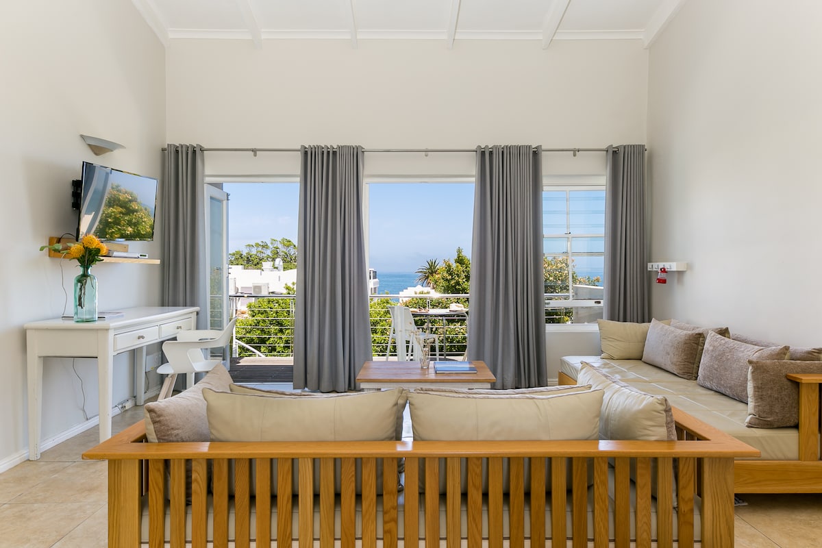 A spacious living area is showcased, featuring a comfortable seating arrangement with neutral-toned cushions. Large glass doors provide a clear view of the outside greenery and the sea, while natural light fills the space. Curtains frame the windows elegantly, complementing the minimalist decor.