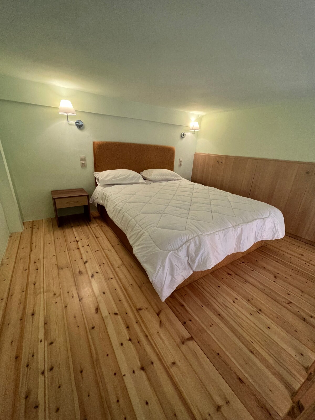 A serene bedroom features a spacious bed with a quilted white cover. Soft lighting is provided by wall-mounted sconces on either side of the headboard. Natural wood flooring contributes to a warm ambiance, with a small bedside table positioned next to the bed.