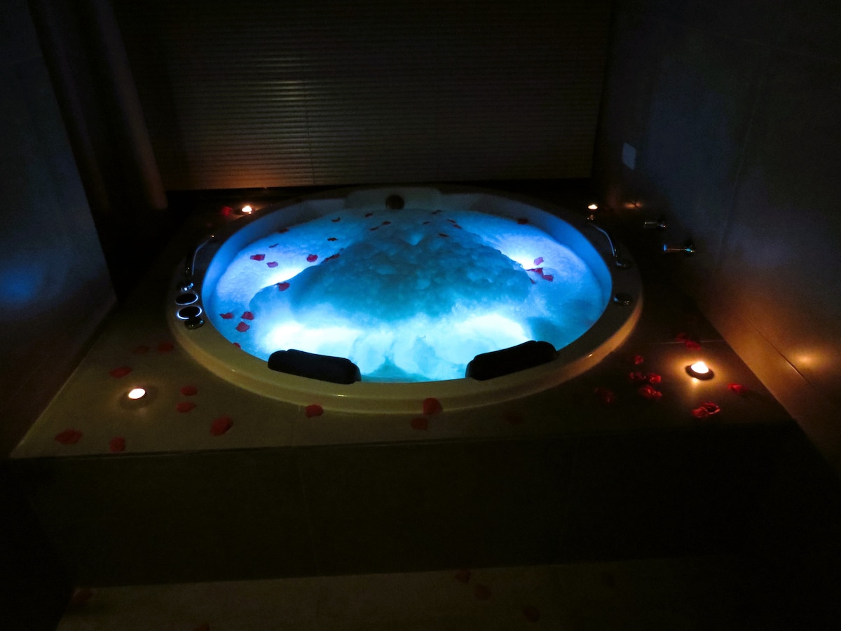 A modern hot tub is situated in a tranquil setting, surrounded by soft blue lighting. Petals are scattered around the edge, enhancing the relaxing atmosphere. Small candles are placed nearby, providing a warm glow to the space.