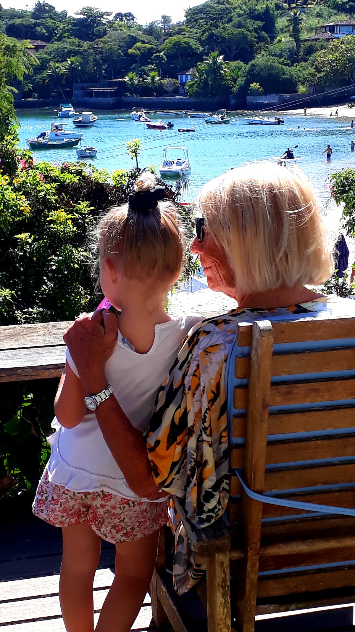 A serene view of a bay is showcased, with boats gently bobbing on the clear blue water. In the foreground, a seated individual enjoys the scene while embracing a child, both overlooking the vibrant greenery and tranquil beach below.