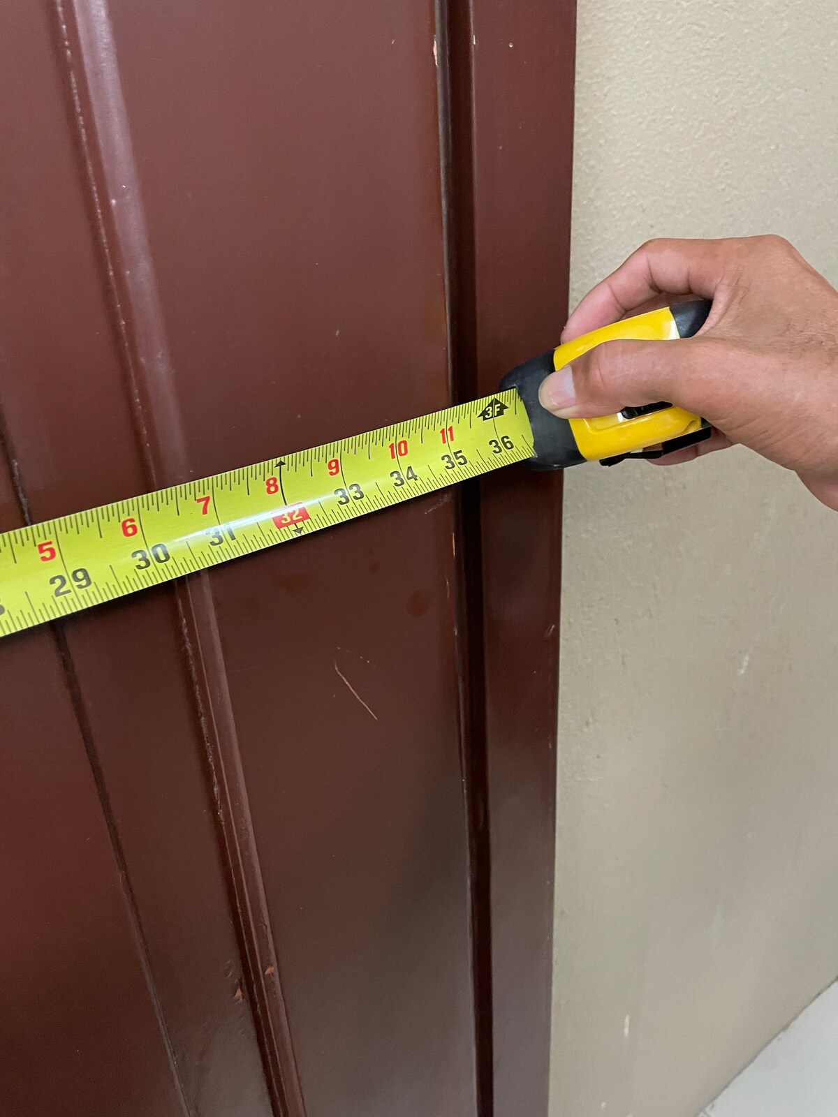 A tape measure is held against a brown door, indicating a measurement along the door's edge. The wall beside the door is a neutral color, and the floor appears smooth and light-colored.