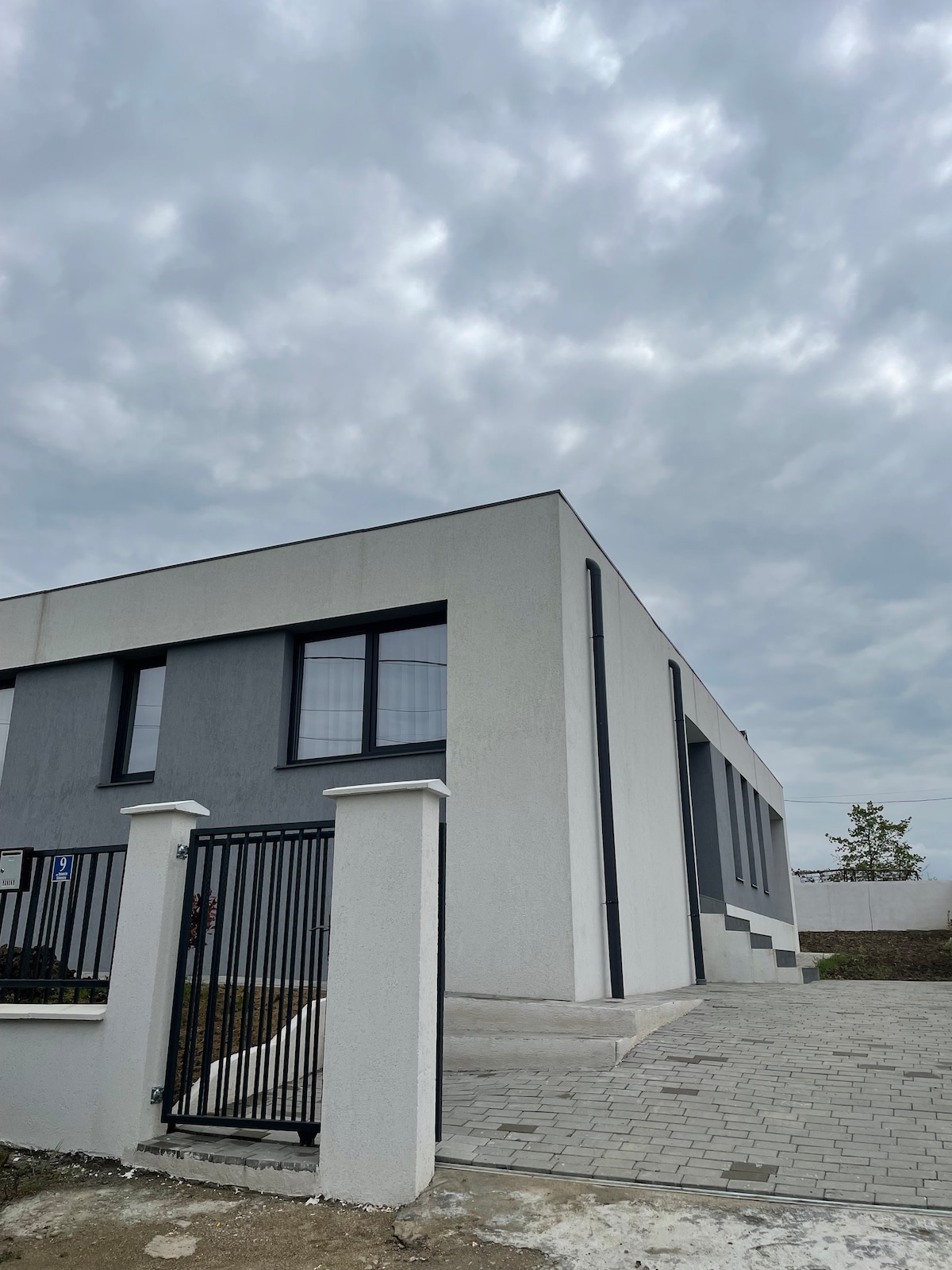 The exterior of the modern building features a mix of gray and white facades. Large windows provide natural light to the interior, and a paved pathway leads to the entrance, which is secured by a black iron gate. Overcast skies create a serene ambiance.