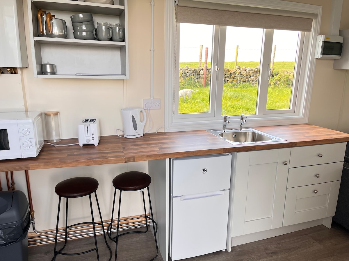 A compact kitchen area features a wooden countertop with a sink, a small refrigerator, a microwave, and a kettle. Two bar stools are positioned beneath the countertop. Natural light enters through a window, offering a view of the surrounding countryside.