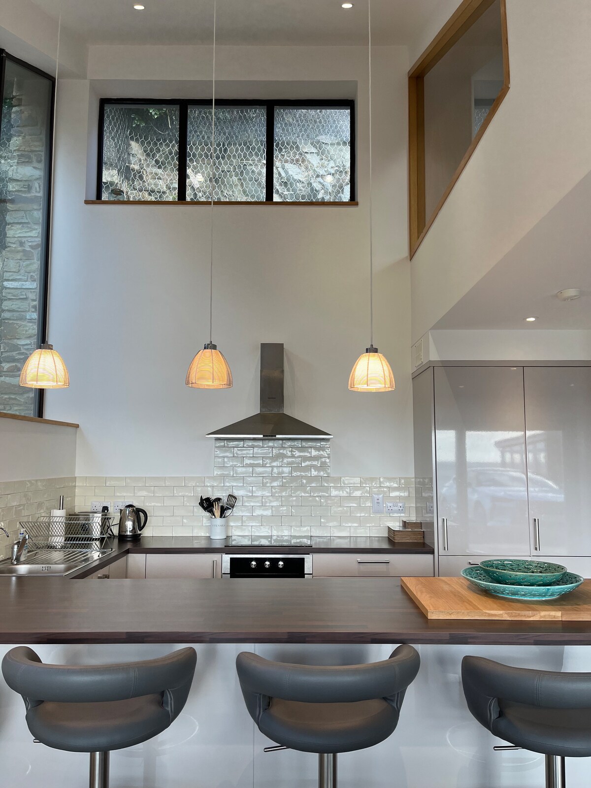 The open-concept kitchen features modern appliances and sleek cabinetry. Pendant lights hang over the countertop, providing soft illumination. Large windows allow natural light to fill the space, while a wooden cutting board and a decorative bowl add subtle touches to the design.