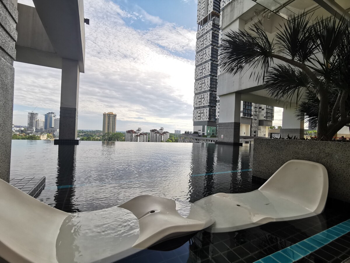 The image showcases a serene infinity pool at a condominium, with comfortable lounge chairs partially submerged. The skyline is visible in the background, framed by modern buildings and lush greenery. Calm waters reflect the sky, creating a tranquil ambiance.