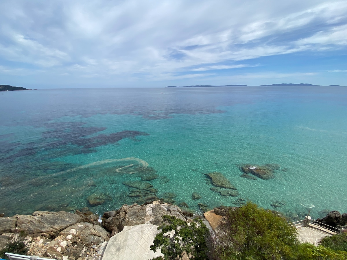 A serene seascape reveals clear turquoise waters extending to the horizon. Rocky formations are visible just beneath the surface, creating a natural contrast with the inner calm of the water. Clouds drift across a light blue sky, enhancing the tranquil coastal setting.