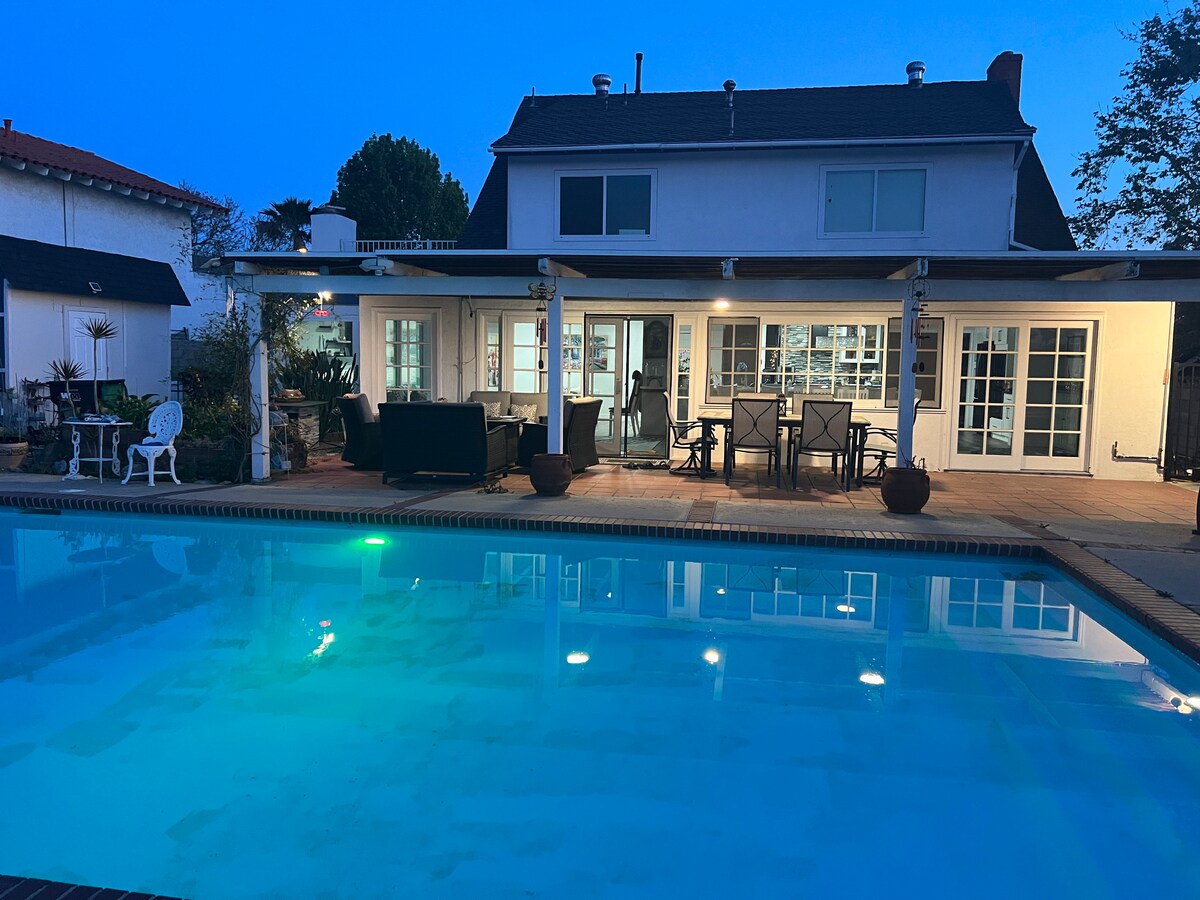 A well-lit outdoor space is visible, highlighting a private pool with submerged lights reflecting in the calm water. Patio furniture is arranged under a covered area, with large glass doors revealing the interior of the house. The pool area is framed by greenery and a clear evening sky.