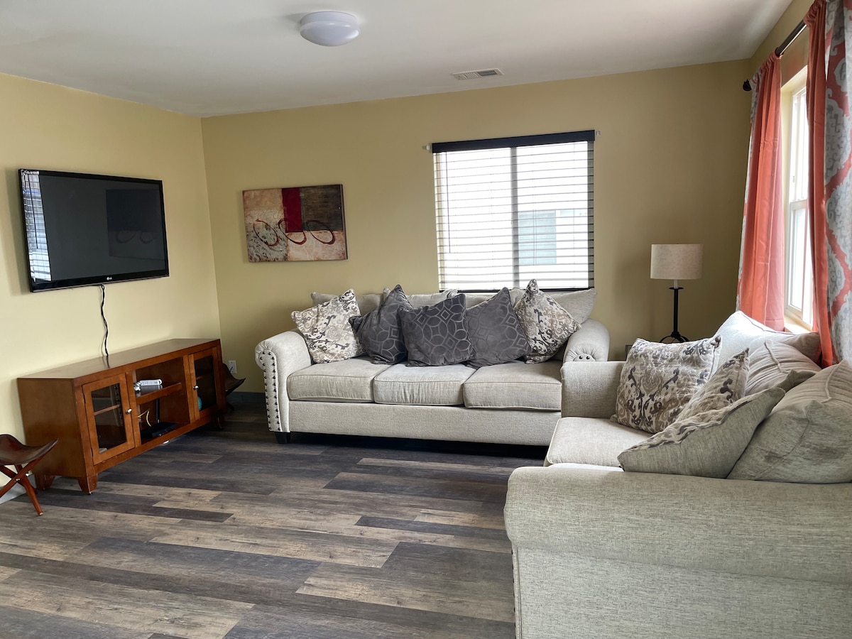 A comfortable living area is shown with a large, light-colored sectional sofa adorned with decorative pillows. A wooden TV stand holds a flat-screen television, while natural light filters through a window adorned with partially open blinds. Warm tones create a welcoming ambiance.