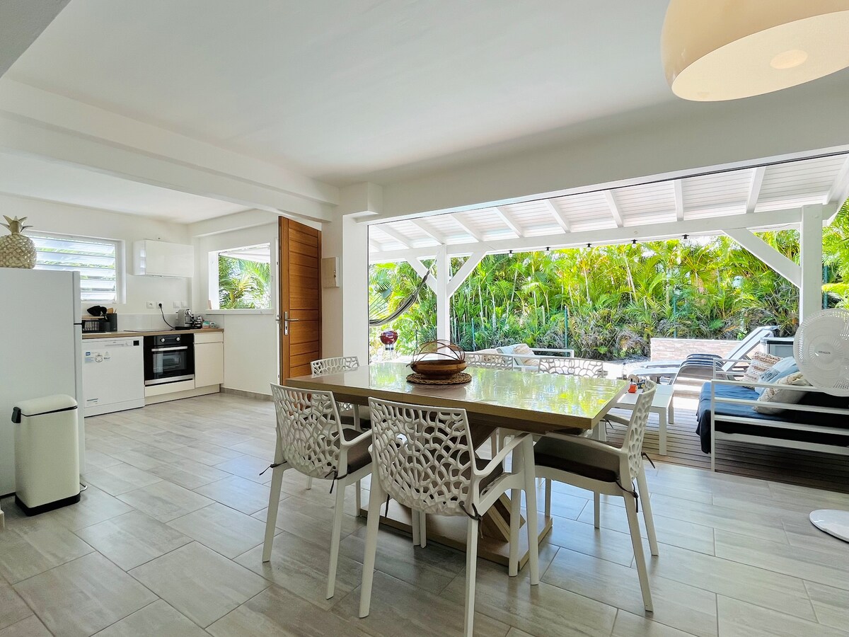 An open-concept dining area with a large table and six white chairs is visible. A modern kitchen is adjacent, featuring white cabinetry and appliances. Floor-to-ceiling glass doors enhance natural light, providing views of lush greenery outside.