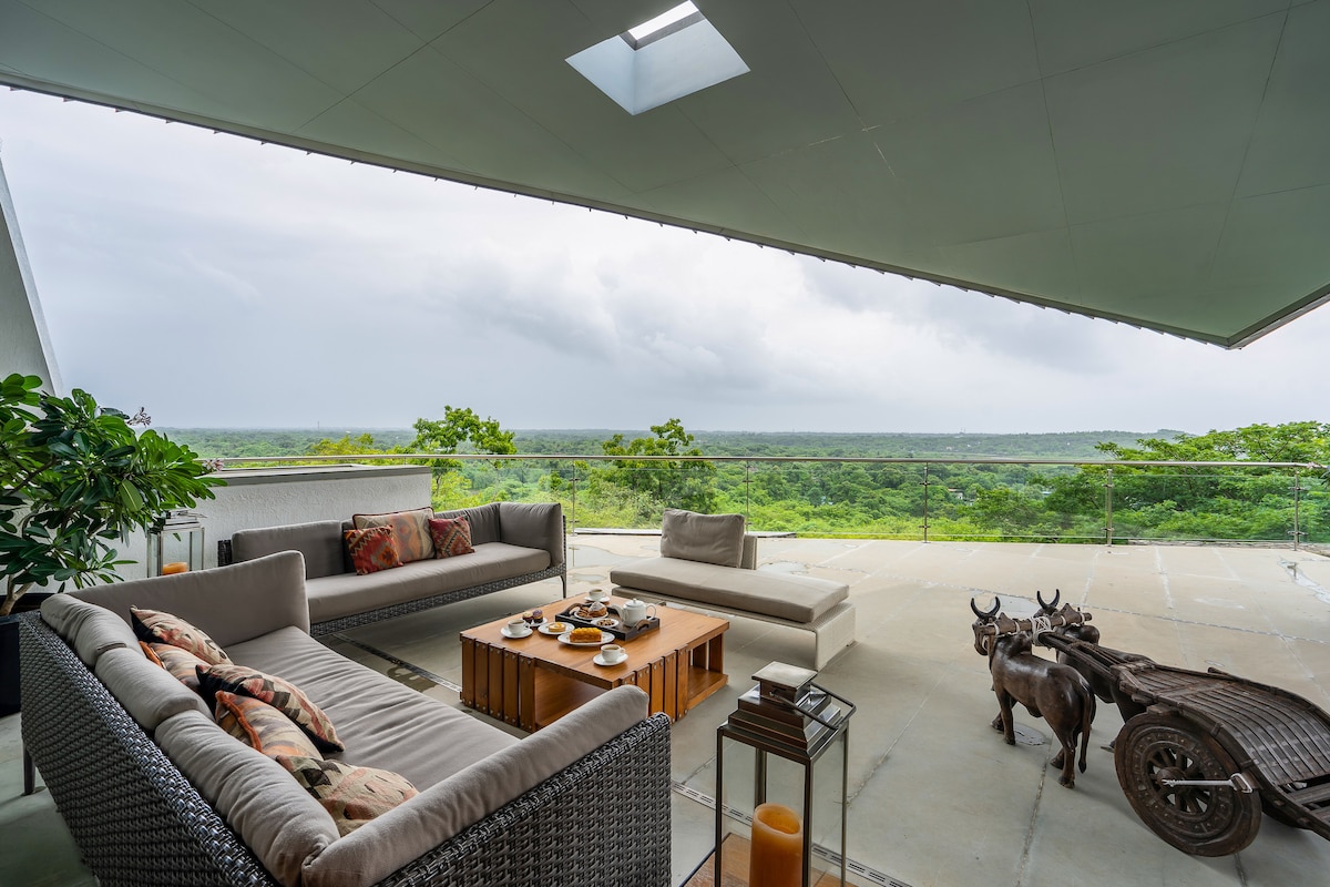A spacious outdoor seating area features comfortable sofas and coffee tables, framed by lush greenery. The area overlooks a panoramic view of the landscape, with a serene atmosphere complemented by decorative items and a traditional cart displayed nearby.