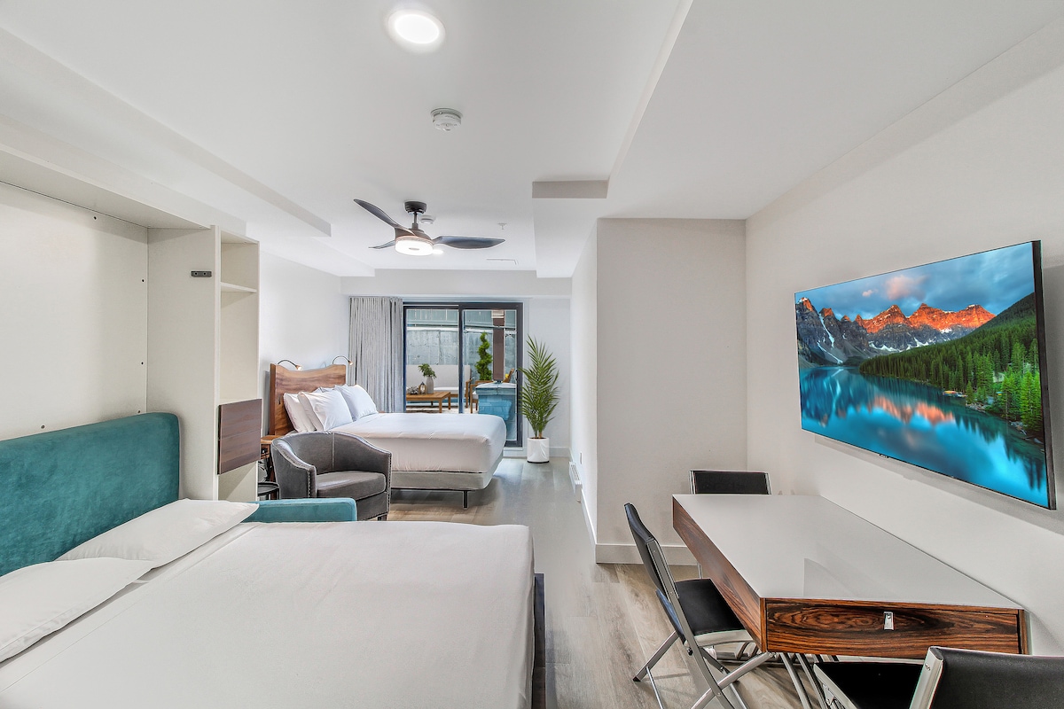 A modern studio layout is displayed, featuring a queen bed against a light-colored wall and a comfortable couch positioned nearby. A dining table is set to the side, with a television mounted on the wall opposite the beds. Natural light enters through patio doors leading to an outdoor space.