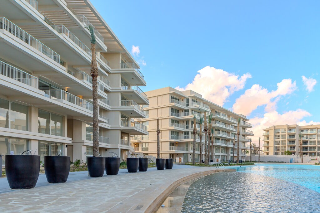 Three modern buildings with expansive balconies are visible along a serene pathway. A calm, crystal-clear lagoon reflects the sky, while large, dark planters line the walkway, adding a touch of greenery to the contemporary design.