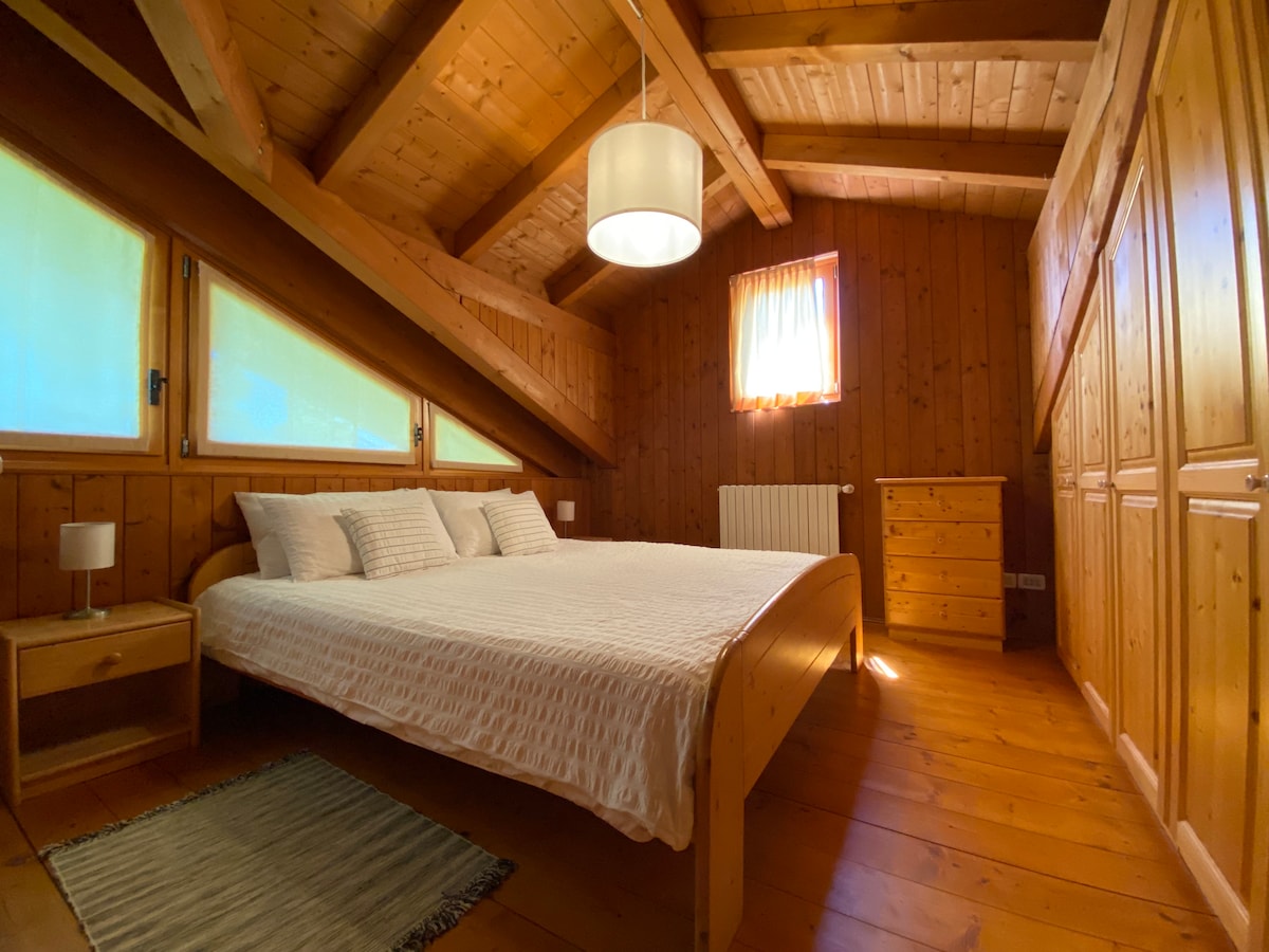 A wooden bedroom features a spacious bed with a light-colored bedspread and decorative pillows. Natural light filters through the windows, illuminating the warm wooden walls and ceiling. A small bedside table with a lamp is positioned next to the bed, and a rug is placed on the wooden floor.