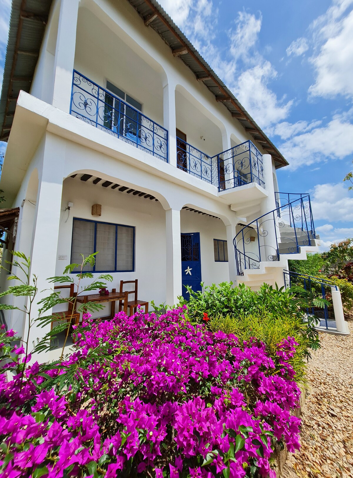A two-story villa features a white exterior with a decorative staircase leading to a balcony adorned with decorative railings. Colorful bougainvillea flowers bloom in front, adding vibrant shades of purple against the backdrop of a clear blue sky and inviting atmosphere.