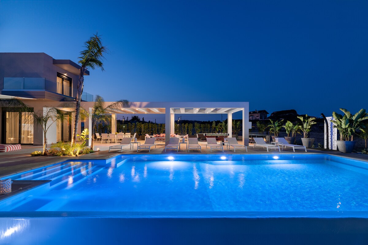 A tranquil outdoor area is showcased at twilight, featuring a spacious pool illuminated in vibrant blue. Comfortable lounge chairs surround the pool, beneath a modern shaded structure. Lush greenery adds a touch of nature, enhancing the relaxing ambiance of the setting.