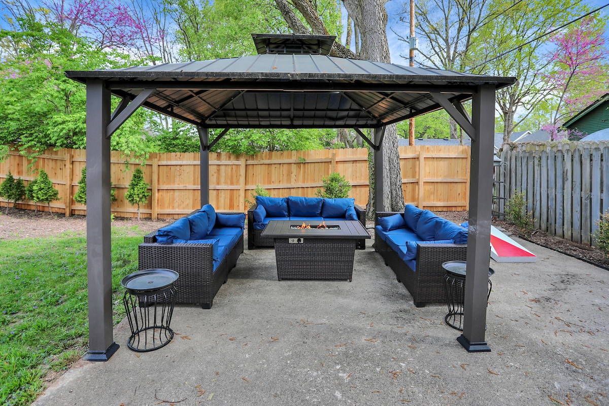 An outdoor seating area is covered by a gazebo, featuring a central fire pit surrounded by dark wicker furniture and blue cushions. Two small tables are positioned next to the seating, with a wooden fence and greenery providing privacy in the background.