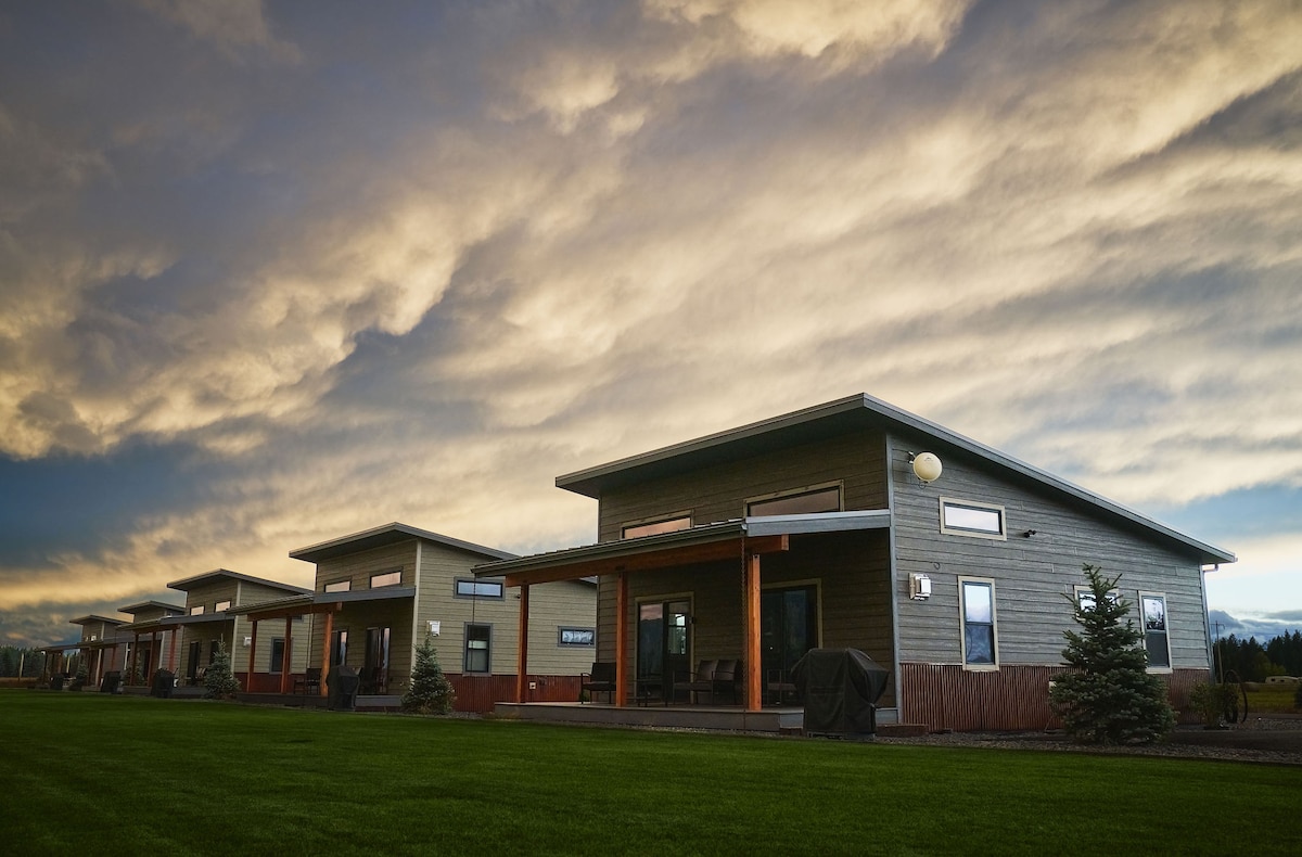 The cabin exterior is showcased against a dramatic sky, featuring multiple structures set on a green lawn. Each cabin is designed with large windows and a welcoming porch, offering a hint of outdoor seating and amenities like gas BBQs.