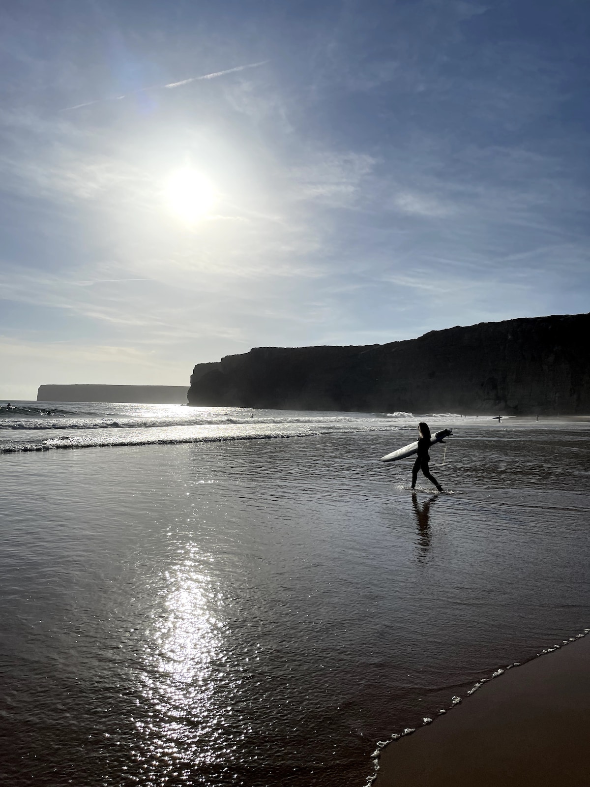 A surfer is seen walking along the shoreline, with a surfboard in hand. The sun shines brightly in the sky, casting reflections on the wet sand. Cliffs rise in the distance, creating a scenic backdrop against the ocean waves.