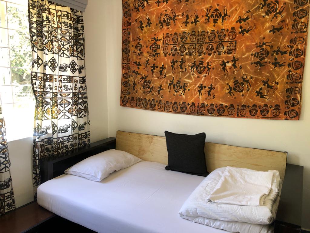 A cozy sleeping area features a single bed with two pillows positioned on a wooden base. A patterned tapestry adorns the wall above the bed. Sunlight filters through a window dressed with patterned curtains, adding a touch of warmth to the space.