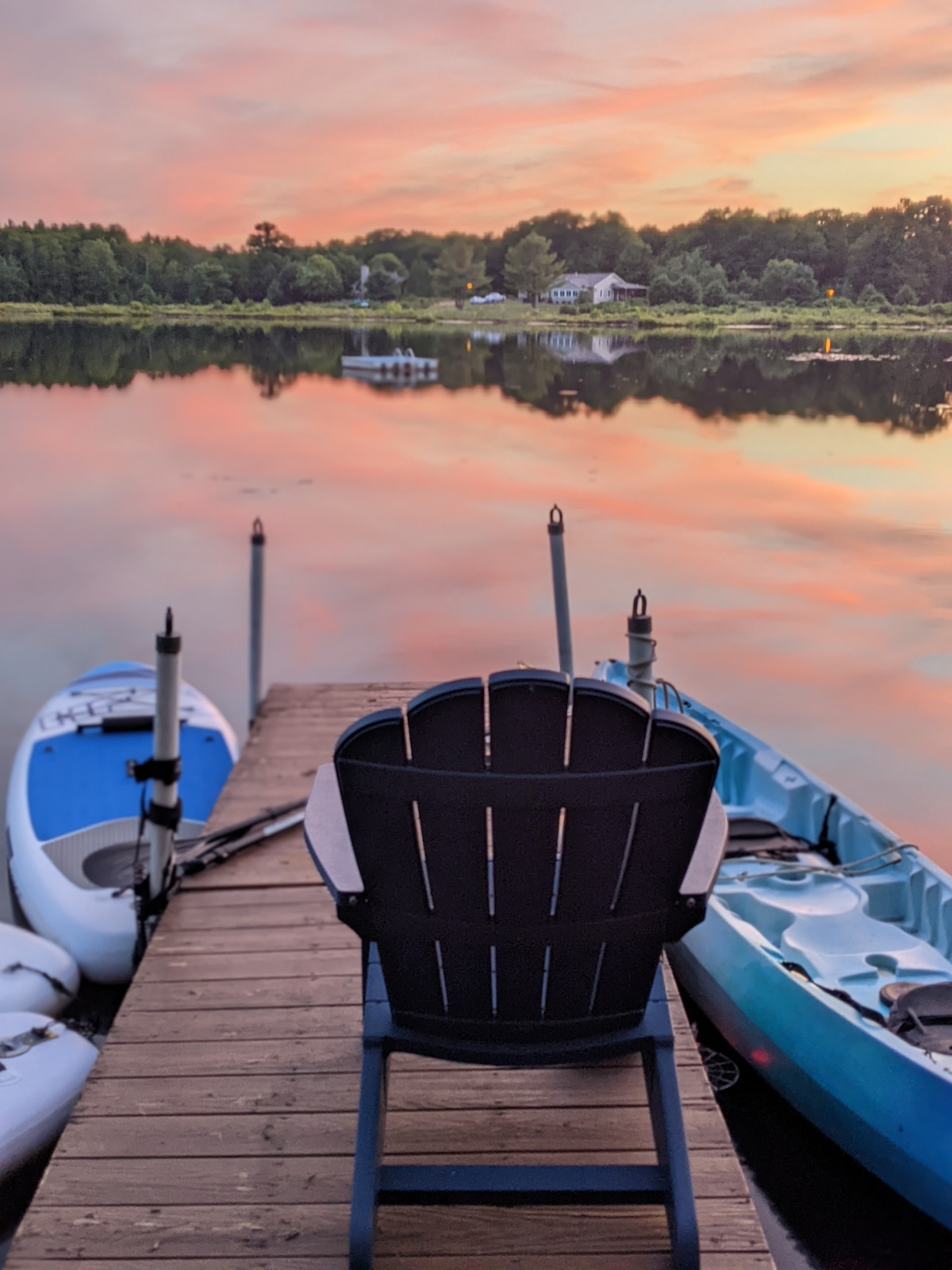 A wooden dock extends over calm waters, flanked by a black Adirondack chair. Two kayaks are parked along the sides, one blue and one white. The scene is bathed in soft sunset hues reflecting on the lake's surface.