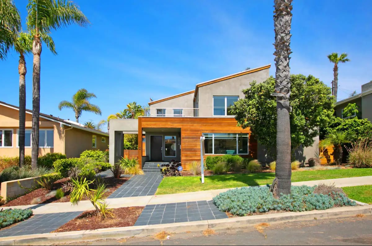 The exterior of the modern beach house is visible, framed by palm trees and lush greenery. A well-maintained pathway leads to the entrance, showcasing a blend of textured materials, including wood and stucco. Clear blue skies add to the inviting atmosphere.