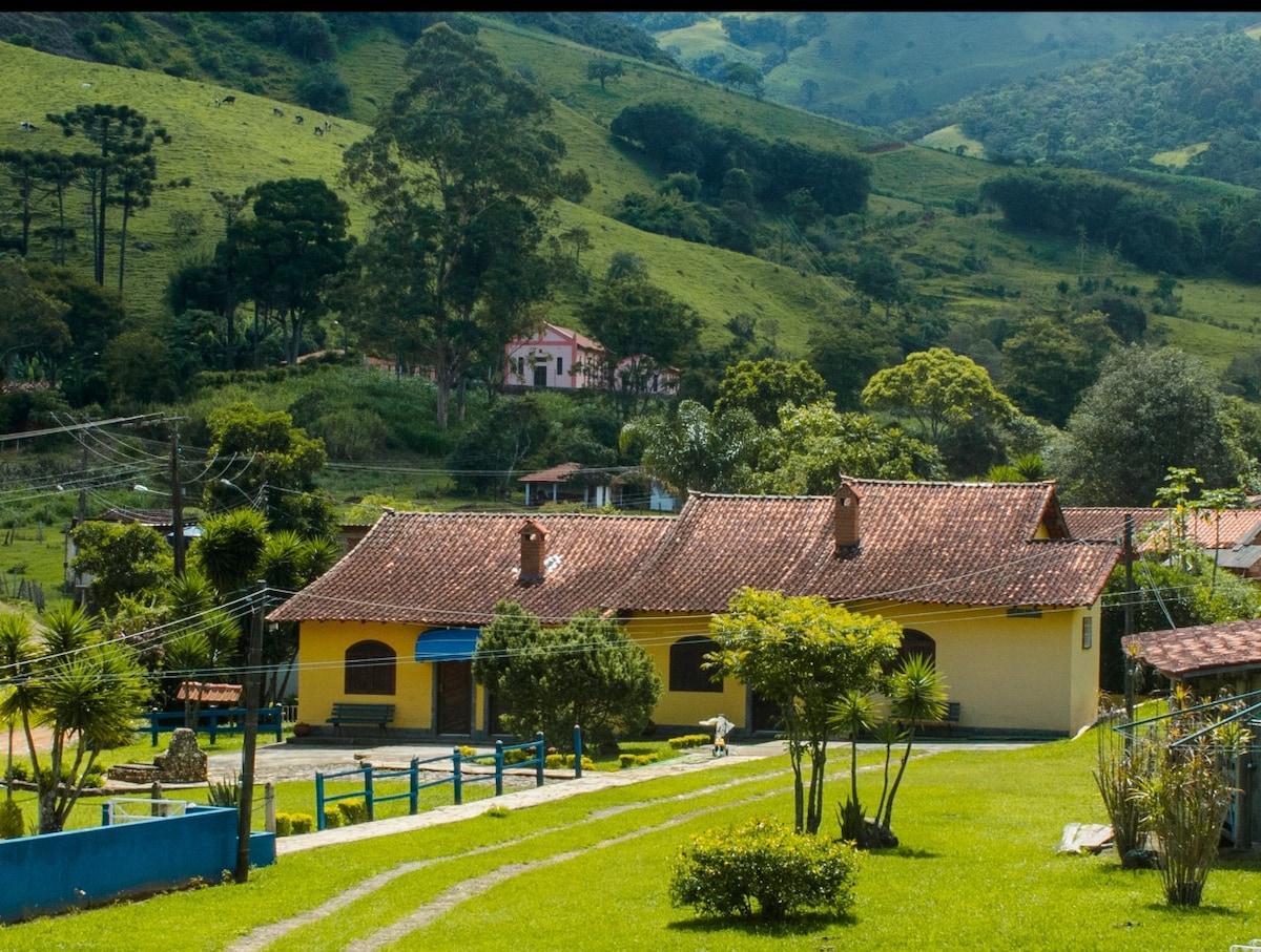 A charming chalet is situated amidst lush greenery and rolling hills. The structure features a traditional tile roof and colored walls, surrounded by well-maintained gardens. Pathways lead to the entrance, showcasing easy access to nature's serene landscape.