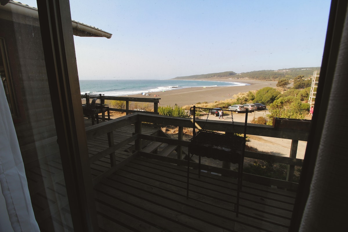 A view from the indoor area showcases a spacious deck leading to a sandy beach. The ocean stretches out in the background, with gentle waves lapping against the shore. Lush greenery and rolling hills frame the picturesque coastline.