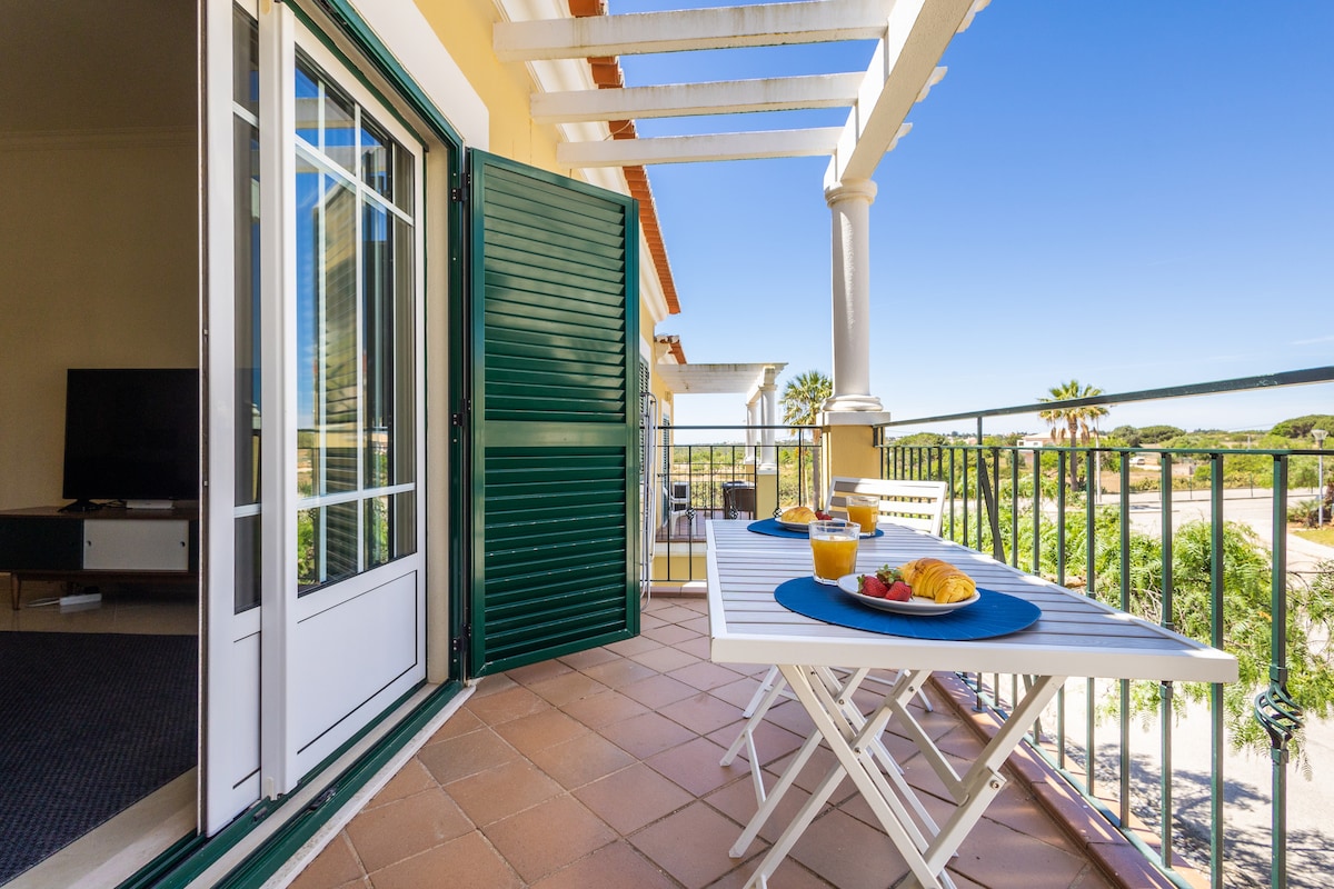 A balcony is shown with two white tables set for breakfast, featuring fruit and beverages. Light filters through the slatted overhang, and a view of palm trees and clear skies is visible in the background. A door leads into the living area, equipped with a television.
